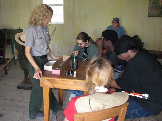 Four young adults seated handling historic pieces of green bottle, park ranger standing explaining artifacts. White walking cane is visible.
