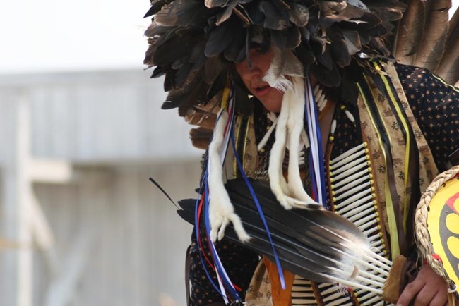 An American Indian dancer wearing traditional dress consisting of a large feathered hat and many beads.