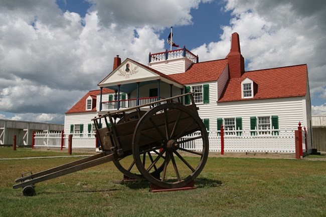 Large brown wooden cart with two large wheels in front of a white house with a red roof and green shutters.