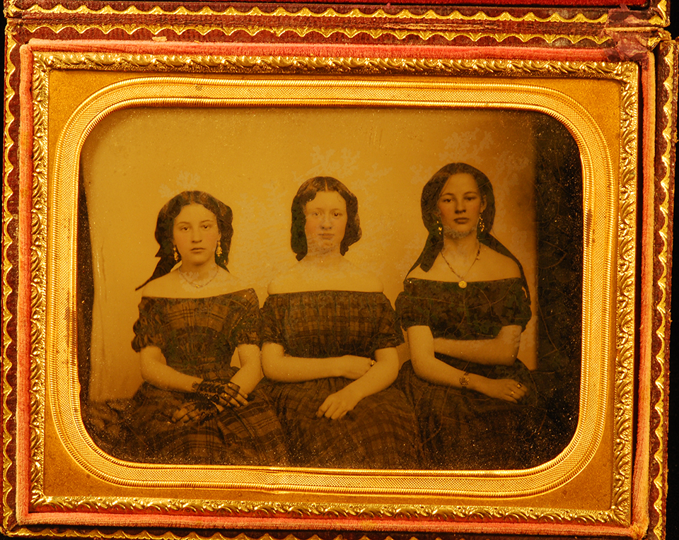 Photo of three girls sitting next to each other wearing 1850s style clothing and jewelry.