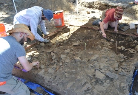 Three students wearing casual clothing use trowels and hand-held brushes to remove dirt from a square of rocky earth.