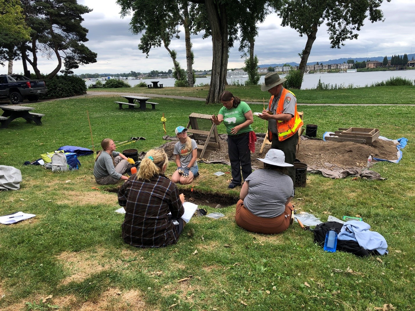 A group of people working on an archaeological excavation in a grassy area by the Columbia River waterfront.