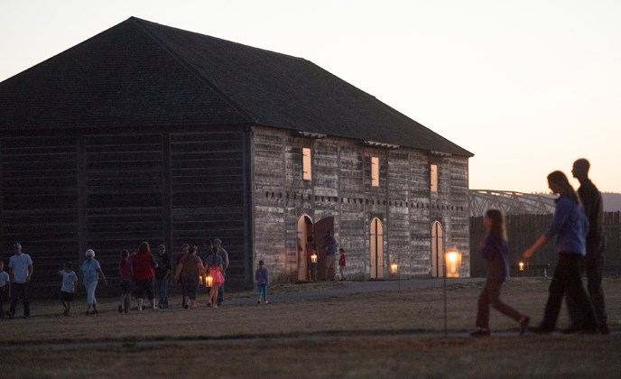 The reconstructed Fur Store at twilight, with candle lanterns outside and people walking around nearby.