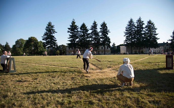 panoramic view of the vintage base ball match game taken behind home plate and showing a hitter swinging at a pitch.