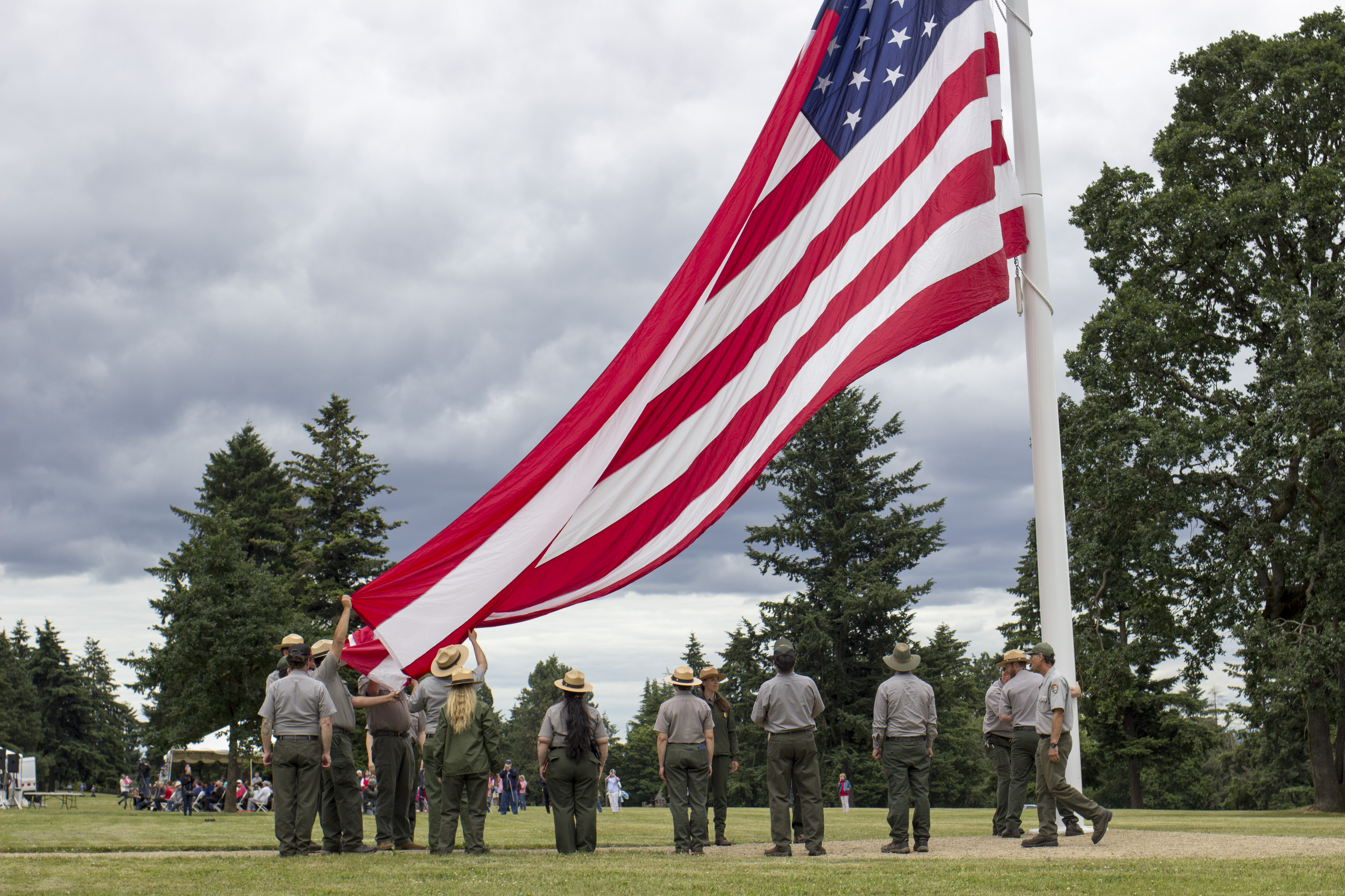 Large flag takes up 3/4 of photo. It is tethered to a pole on the right, and swoops downward to left. Park Rangers hold the bottom of the flag.