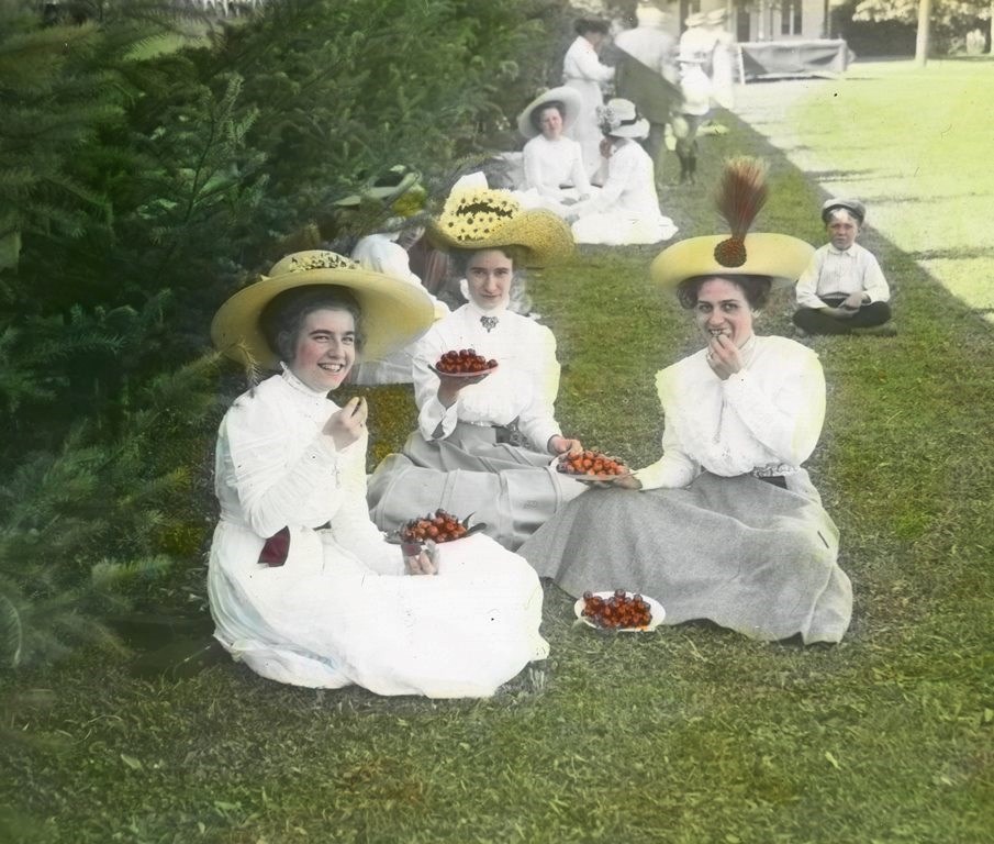 Historic lantern slide image of women wearing light colored dresses and straw hats sitting on lawn eating berries.