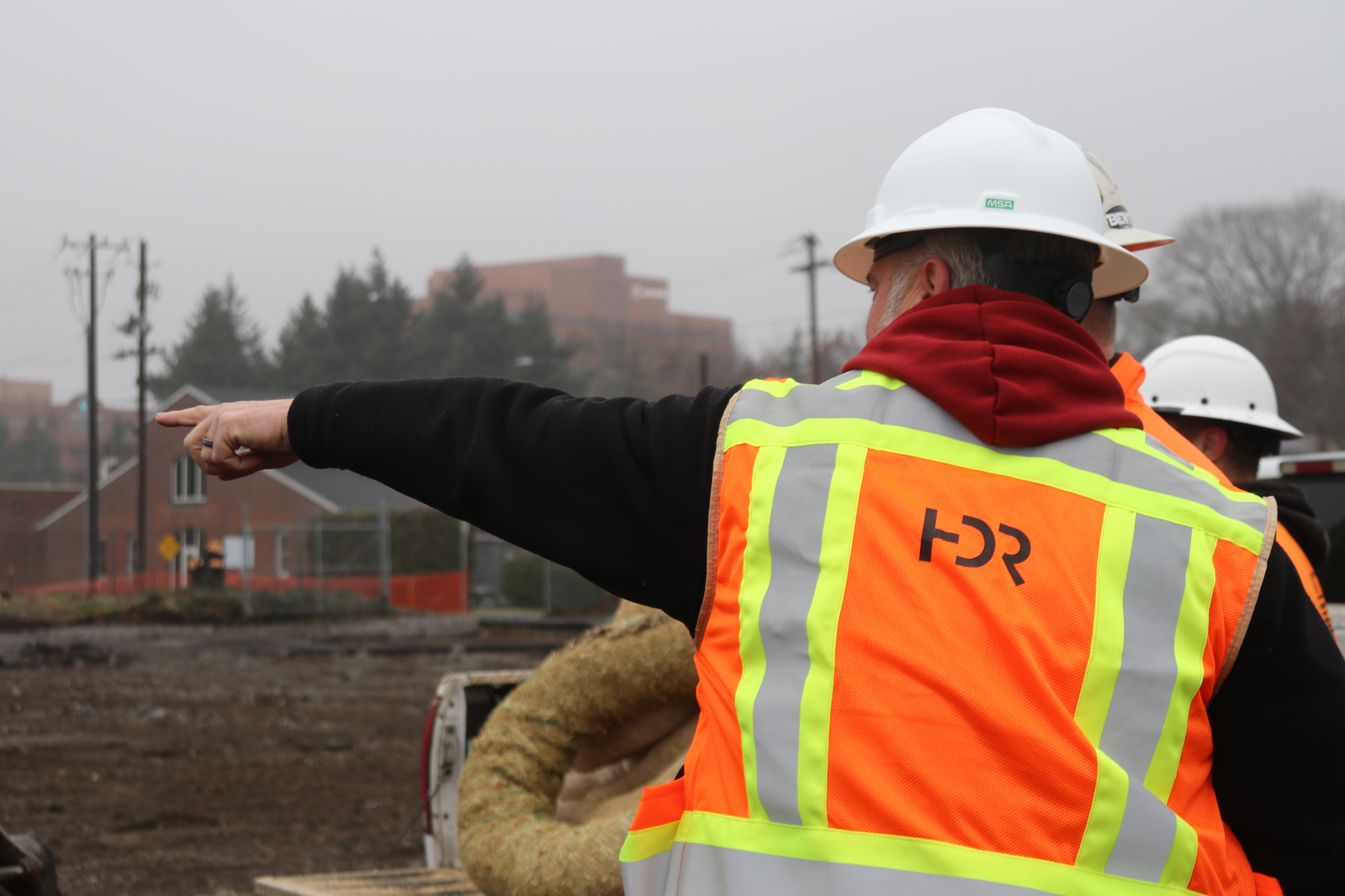 In foreground, man wearing orange safety vest and hard hat points to the distance. Outdoors.