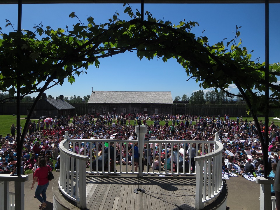 Photo taken from the Chief Factor's House veranda of a large amount of children sitting on the field in front of the house.