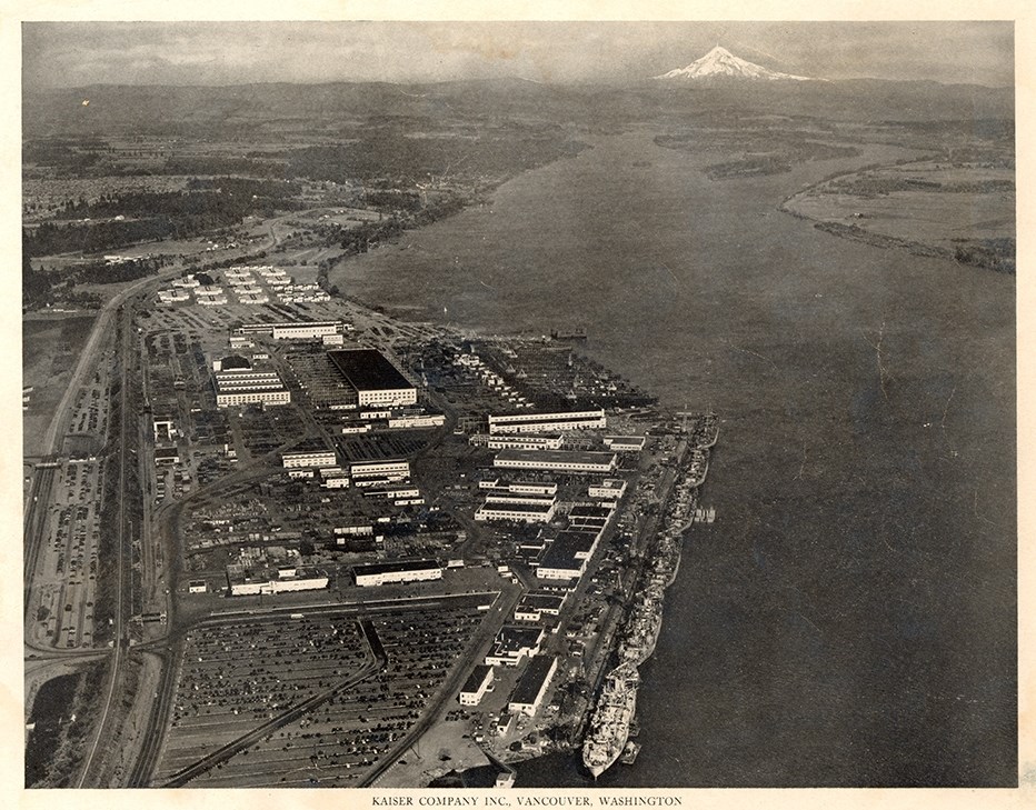 Aerial photograph of the Vancouver Kaiser Shipyards on the Columbia River. Six Navy ships are docked. Mount Hood is visible in the background.