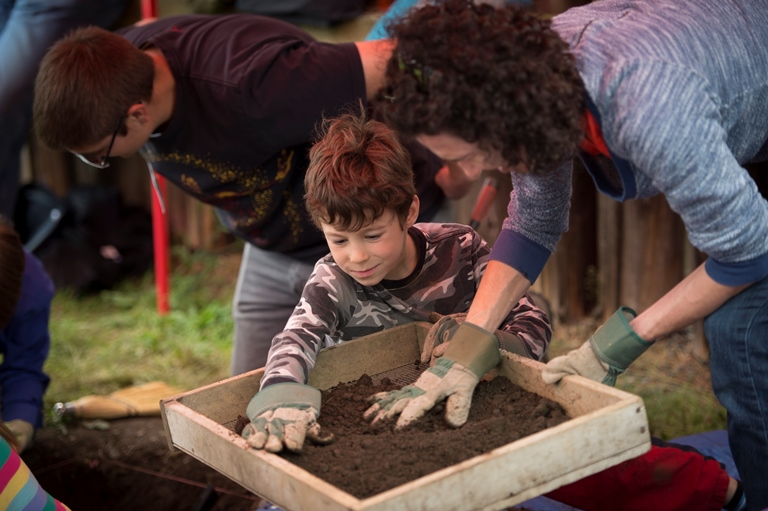 Kids-Dig-for-web.jpg A boy wearing thick gloves sifts dirt through an archaeological screen with the help of a woman who stands behind him.