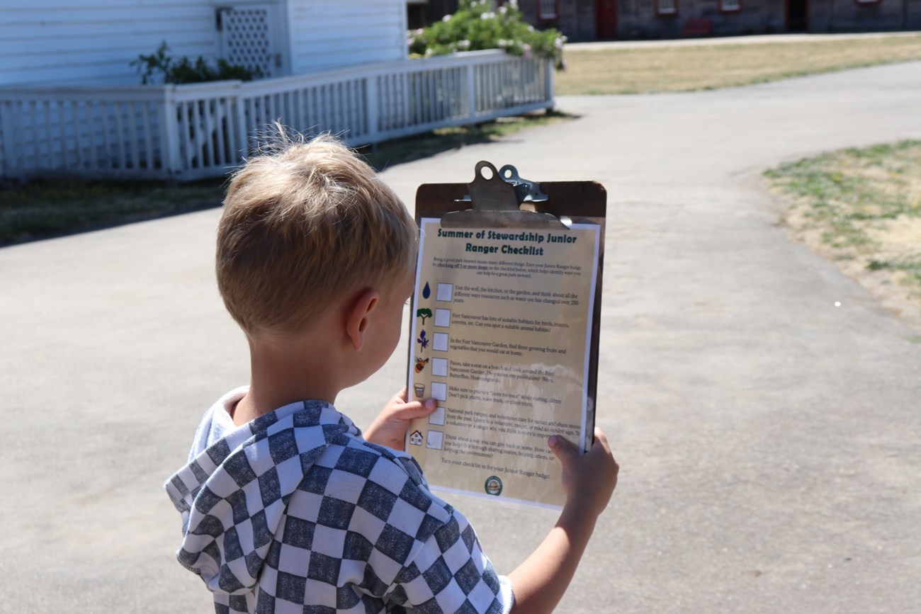Summer Junior Ranger A young child holds up a clipboard with a Summer Stewardship Junior Ranger Checklist inside Fort Vancouver on a sunny summer day.