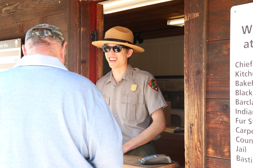 A national park ranger wearing sunglasses on a sunny day smiles as he welcomes a visitor to the Fort Vancouver Contact Station.