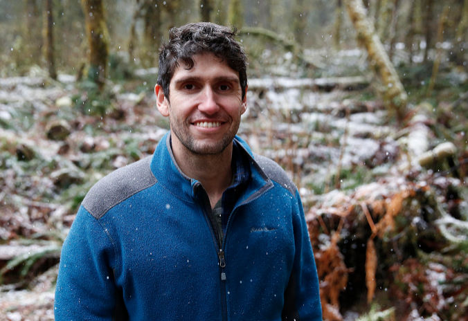 Photo of a man wearing a blue sweater standing in a forest.