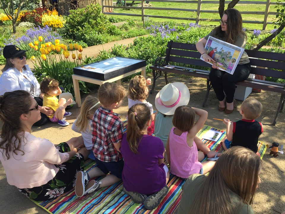 Photo of children sitting in a garden listening to an adult reading a picture book.
