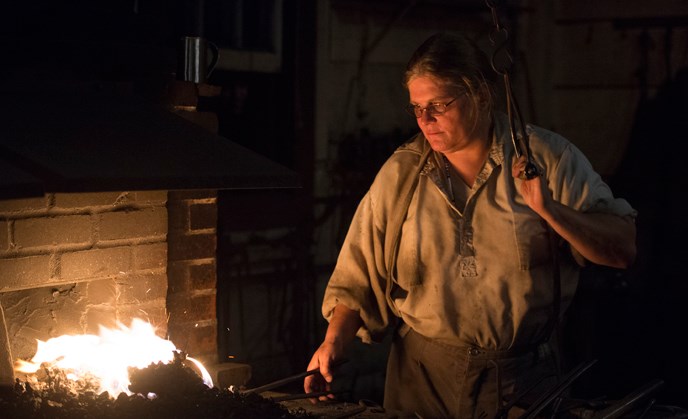 A blacksmith shop volunteer in 1840s costume works a hot forge during a nighttime special event. A blacksmith shop volunteer in 1840s costume works a hot forge during a nighttime special event.