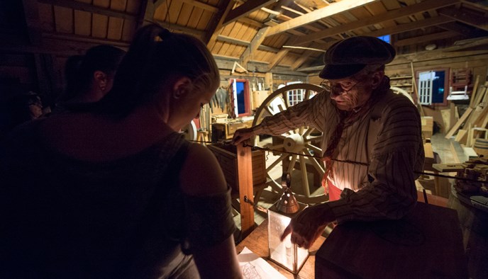 Photograph of a volunteer carpenter dressed in an 1840s work shirt and hat speaking to a female visitor inside the Carpenter Shop during a nighttime special event. Photograph of a volunteer carpenter dressed in an 1840s work shirt and hat speaking to a female visitor inside the Carpenter Shop during a nighttime special event.