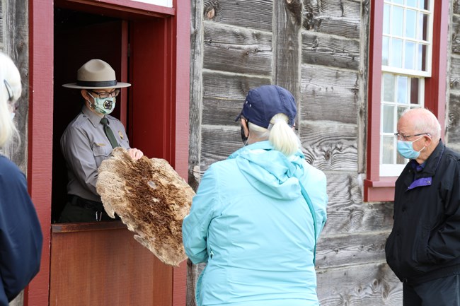 Female park ranger standing in narrow doorway while holding a round beaver pelt. Visitors are feeling the beaver pelt as the ranger speaks.