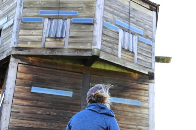 Woman in foreground looks up at large wooden bastion tower with three square windows on the side, well above eye level.