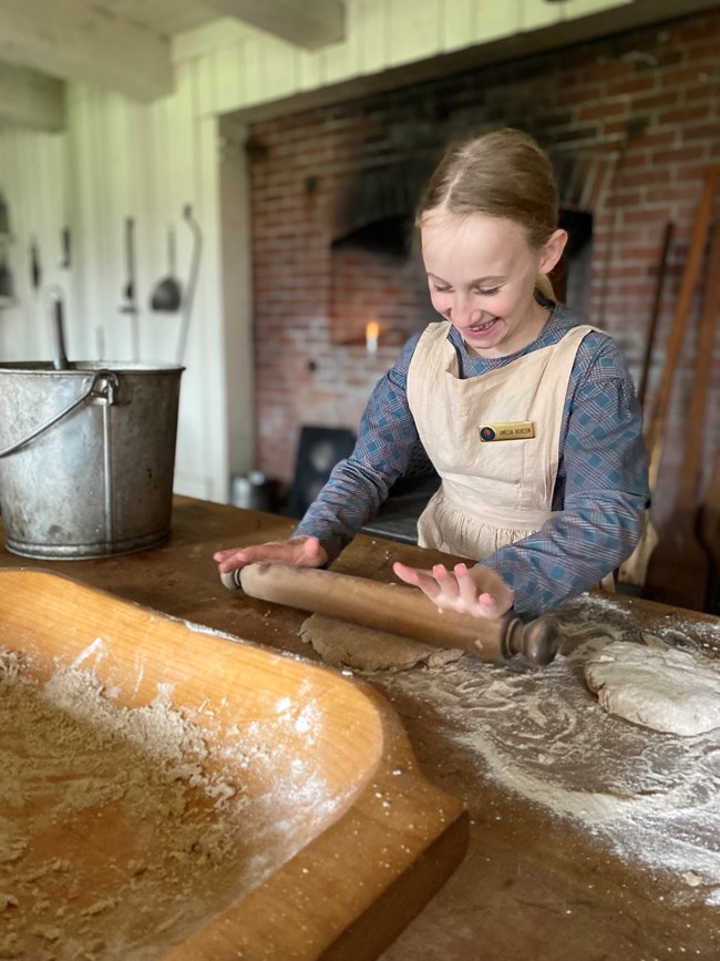 Young blonde girl in historic costume dress uses rolling pin on dough.