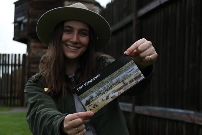 Landscape oriented photo of smiling park ranger holding park brochure with wooden bastion in background.