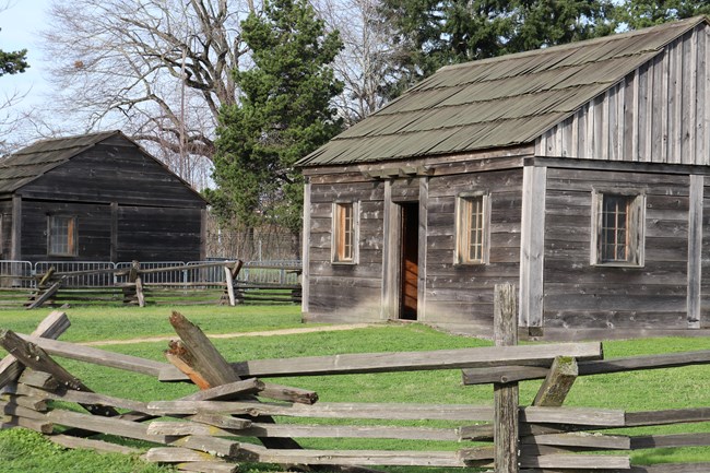 Two wooden one-room cabins stand in a field of green grass with a rustic, stacked, wooden fence in the foreground