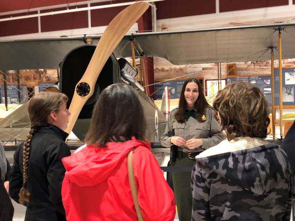 A woman in a park ranger uniform talks to people in front of a Curtiss Jenny aircraft.