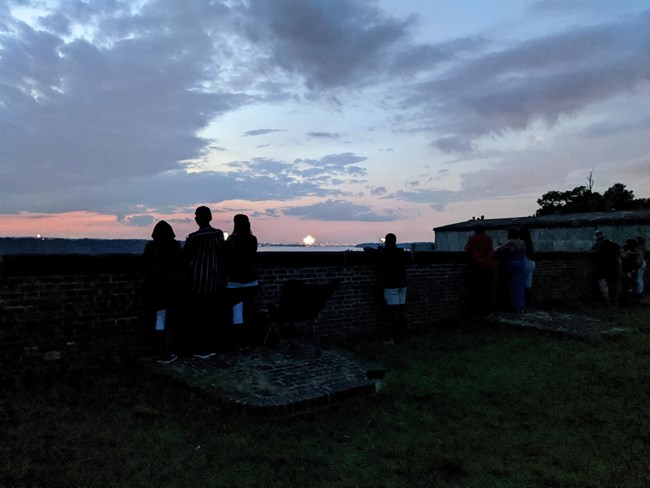 three people stand at the fort wall at dusk watching fireworks down river over the DC skyline