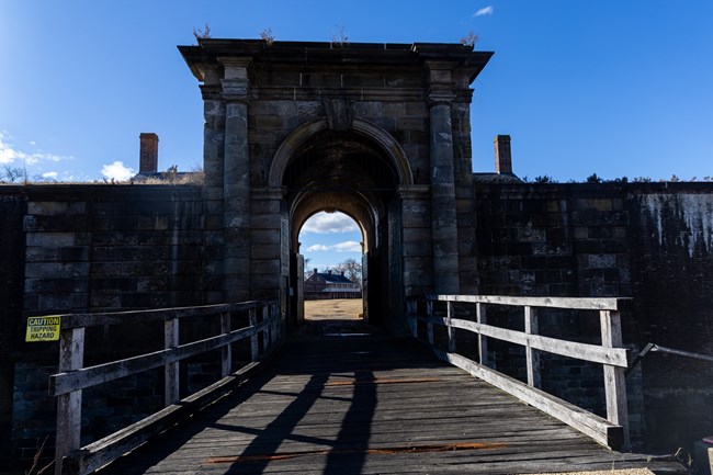 A weathered wooden bridge with railings leads to a tall stone archway through a stone wall.