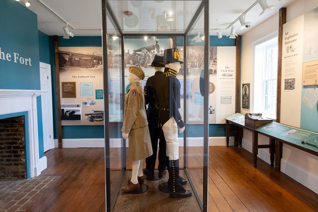 Mannequins dressed in colonial era clothing inside a tall glass case in the middle of a room with informational exhibits on the walls.