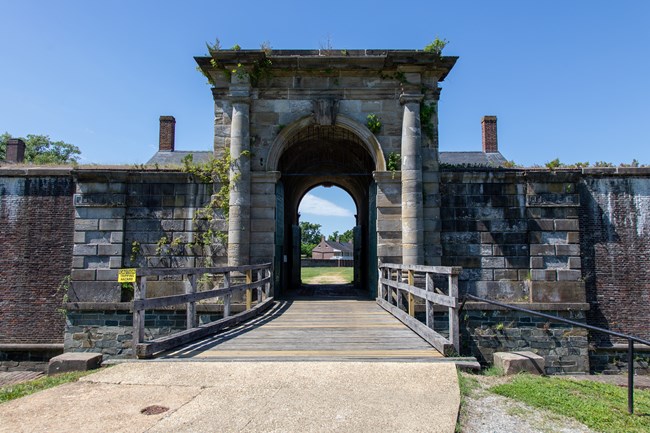 A wood bridge leads to an arched entrance through a tall stone fort wall.