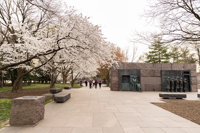 The entrance to the 2nd term room is wide and granite. Trashcans and benches are positioned to the left. Various statues are seen in the background to the right. Cherry blossoms stand to the left.
