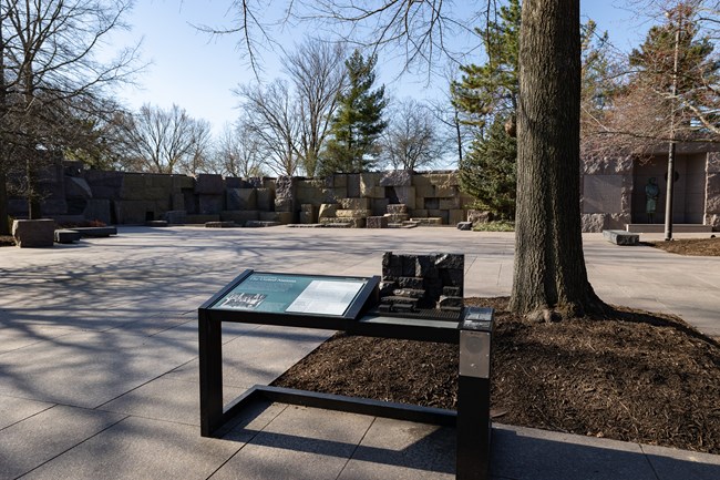 An information panel is positioned in front of a rock formation. To the right is a statue of Eleanor Roosevelt wearing a dress and standing tall. There is a miniature, tactile version of the rock formation on the information panel.
