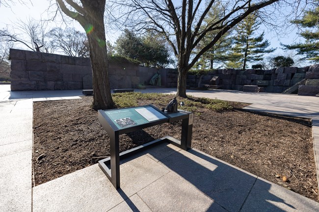 An information panel is positioned in front of a statue of FDR and a statue of a dog. In between the panel and the statues is a tree with dirt surrounding it. To the right is a rock formation and fountain.