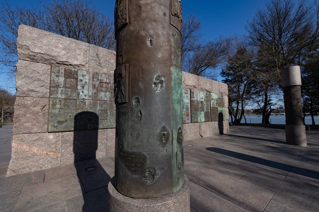 A bronze column has various tactile elements displayed such as braille and human faces. Tiles behind the column also have similar tactile elements displayed.