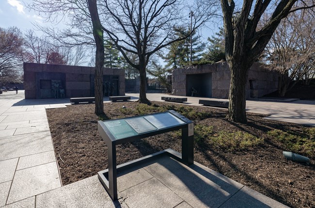 An information panel is positioned in front of various statues in the 2nd term room. In between the statues and information panel is a planted tree surrounded by dirt.