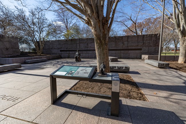 An information panel is positioned in front of a life-sized statue of FDR in a wheelchair. On the information panel is a miniature, tactile version of the statue, and audio description.