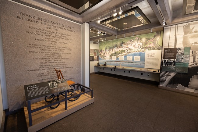 A life size wheelchair is positioned to the left with an information panel in front of it. To the left sits a long table with various exhibits that include audio, braille, and tactile elements.