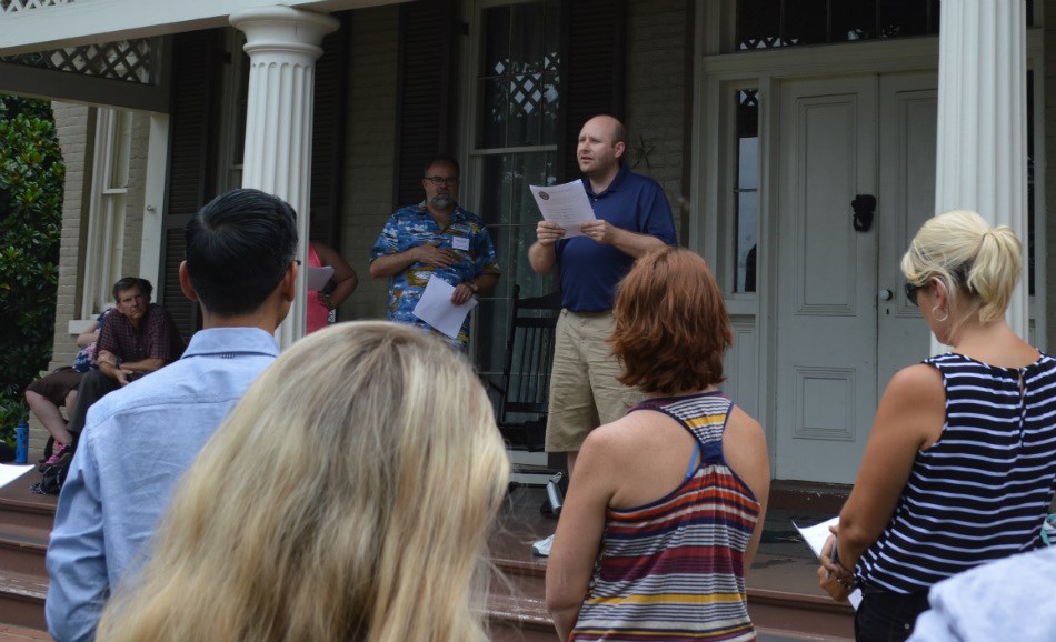 A man stands on a porch and reads from a paper to an audience