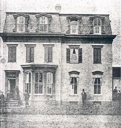 lt= A black-and-white photograph of Frederick Douglass standing in front of a home