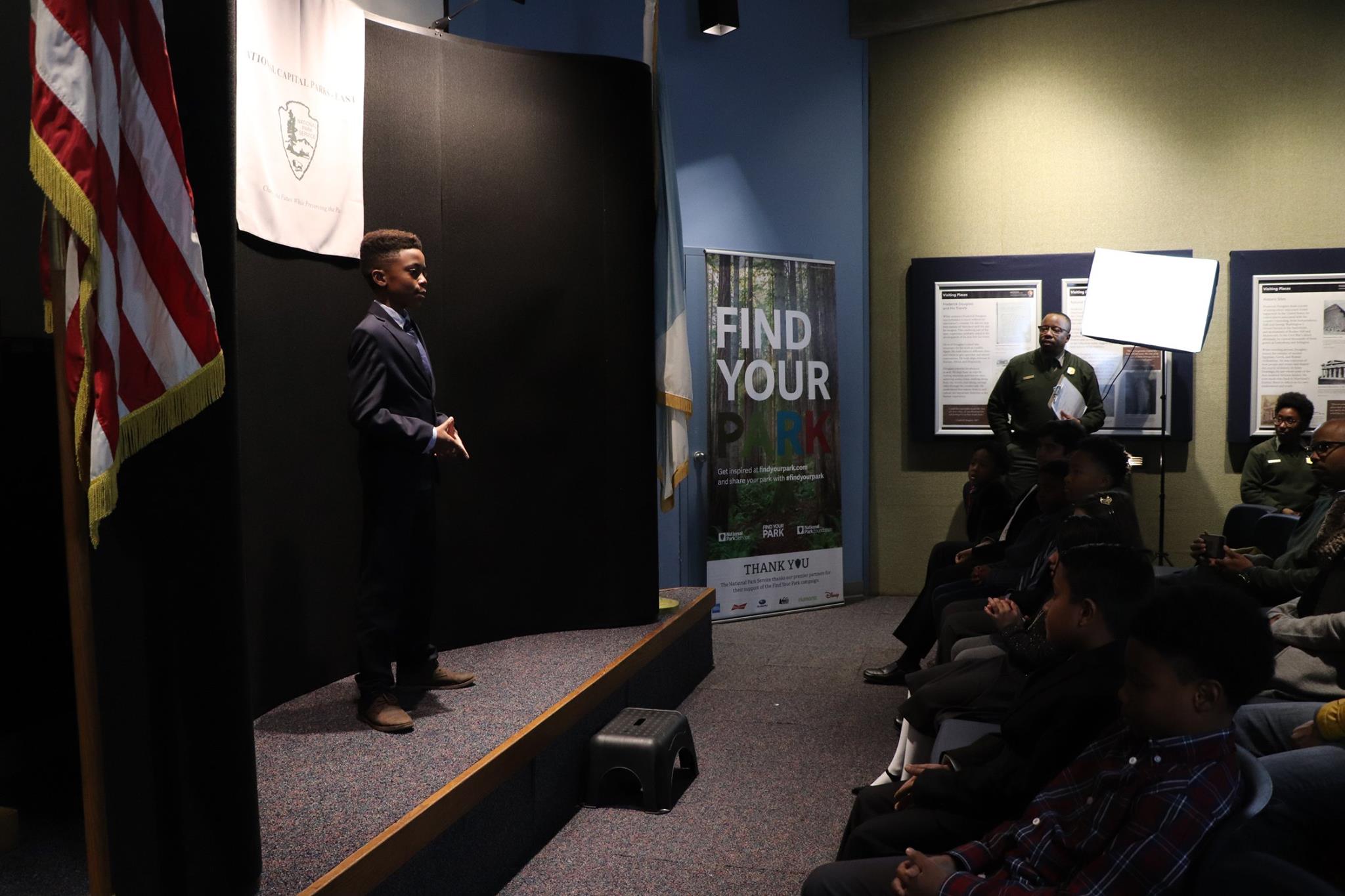 A student stands on a stage and gives a speech to a small audience