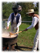 Two men in historic costume use wooden tools to stir soup in a cast iron kettle.