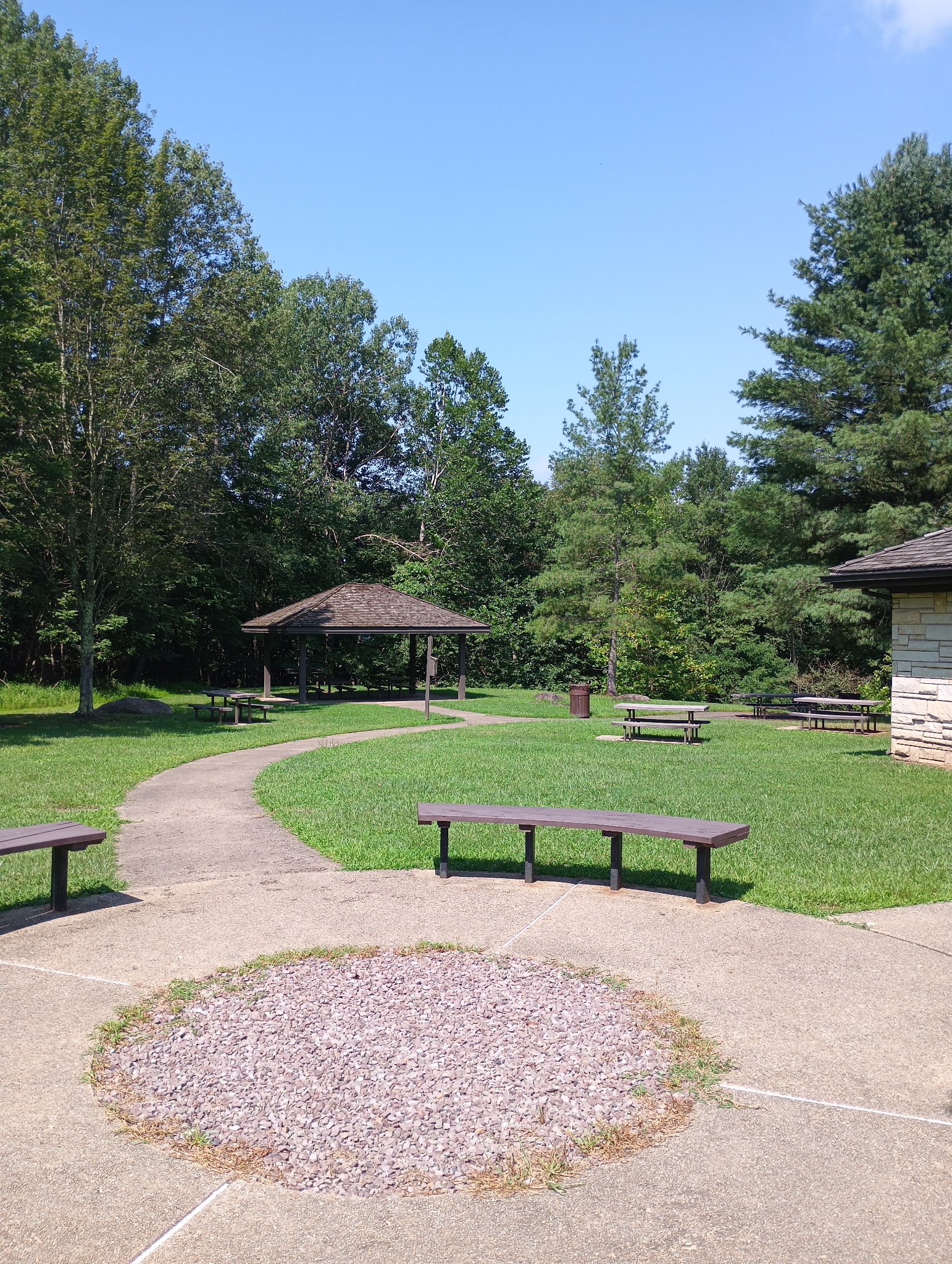 Grassy area with concrete sidewalks and a couple benches. A corner of a building on the right. Pavilion and picnic tables backed by a bunch of trees in a scenic picnic area for permitted weddings.