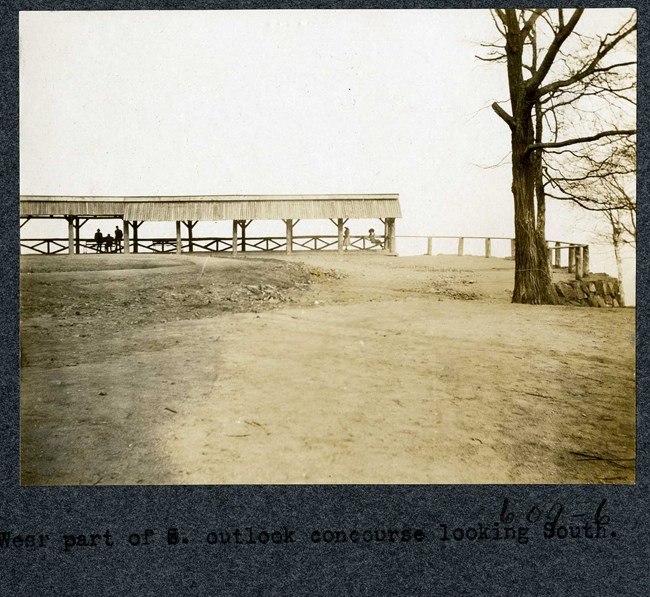 Black and white photograph of top of mountain, with wooden railing on the edges and a wooden shelter, with a few people standing under it