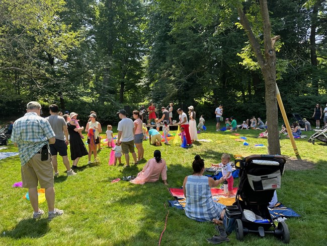 Group of children and adults sit and stand on flat grassy area with trees and stage in front