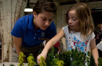 Woman in blue shirt bends down next to girl placing miniature trees in model