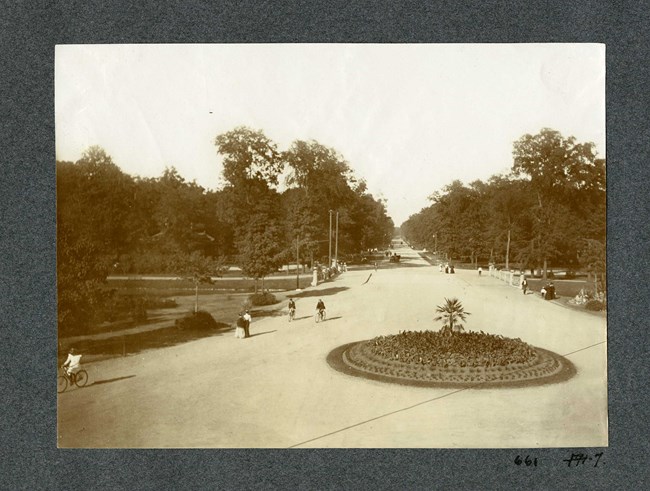 Black and white photograph of long pathway cutting though a park, with large open green areas on each side with some trees lining them. On the path people walk and bike, and there is a circular flower display in the middle.