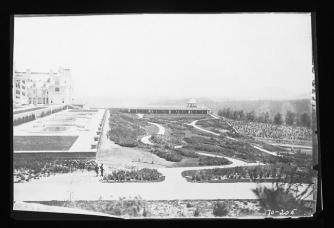 Black and white photograph of hillside garden in front of a large mansion. The garden in front is rectangular with many curved rock paths cutting through it