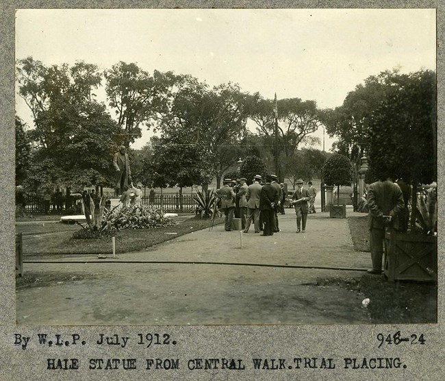 black and white photograph of men in suits and hats walking along a dirt path lined with patches of grass. One patch contains many plantings and a statue.