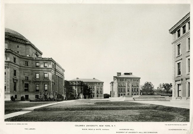 Black and white photograph of buildings all surrounding a rectangular patch of grass. The grass is surrounded by dirt walking paths