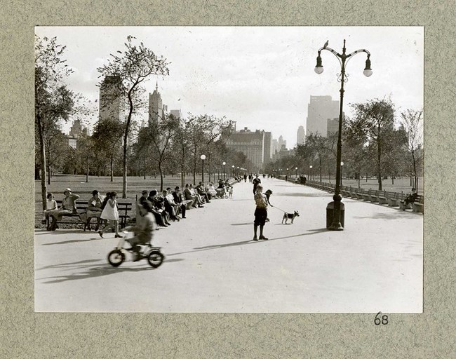 Black and white photograph of rectagular dirt path cutting in half two large green spaces. The dirt path is lined with benches, many of which are filled, as some ride bikes or walk with dogs in the center. On the grassy side, trees line the walkway and in